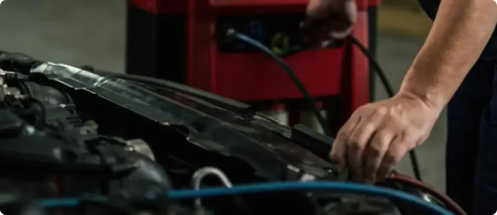 Close-up of a technician's hands working on a vehicle's engine bay connected to professional red diagnostic equipment with blue hoses, likely for AC or system testing.
