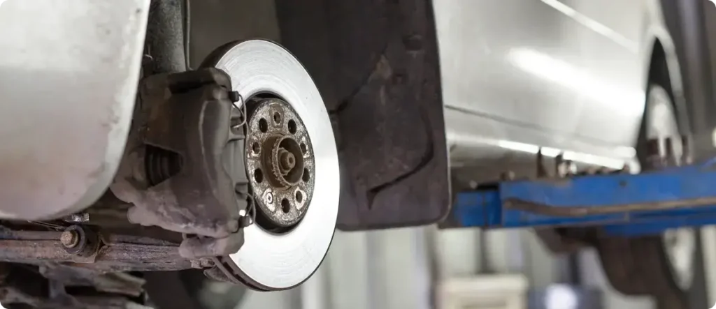 Close-up of a car brake disc and caliper with the wheel removed, showing brake system components during vehicle maintenance on a lift.