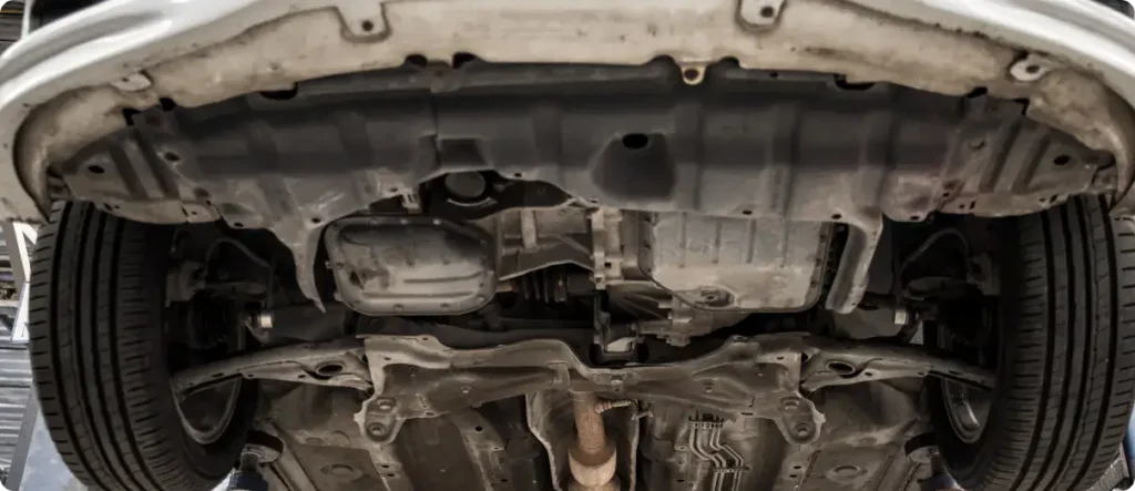 Underside view of a car on a lift showing suspension, exhaust, and chassis components during a comprehensive vehicle inspection in an auto repair shop.
