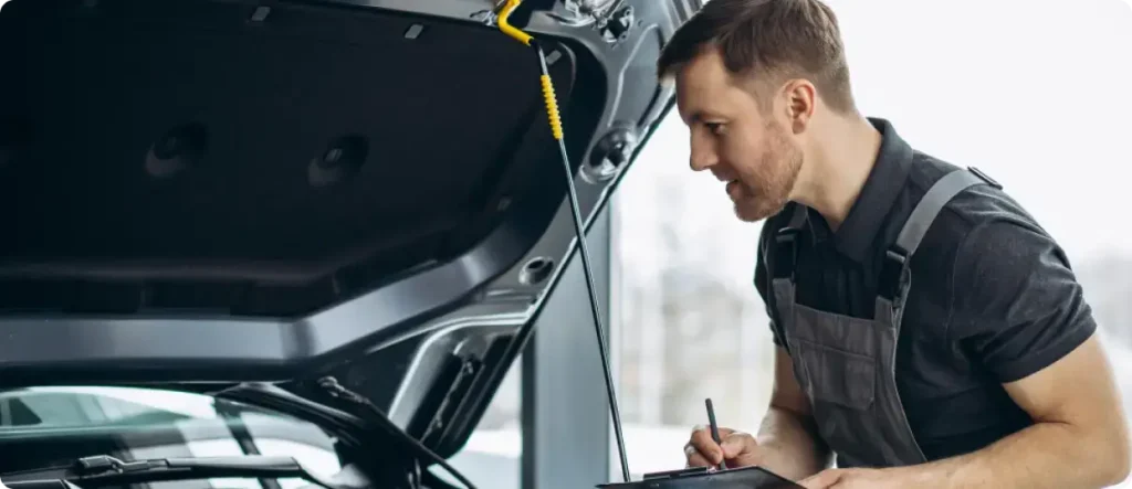 A technician in grey work overalls standing beside an open car hood, carefully filling out a multi-point inspection checklist on a clipboard.