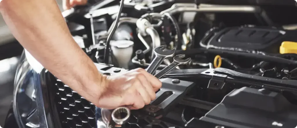 Mechanic holding a wrench over an open car engine bay, preparing to perform maintenance or repair inside a professional auto service garage.