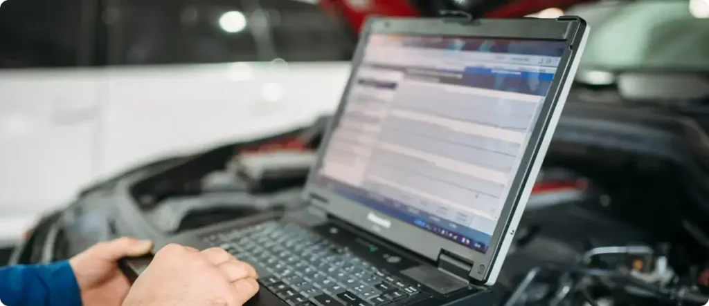 Mechanic using a diagnostic laptop connected to a vehicle in an auto repair shop, analyzing car system data during professional maintenance.