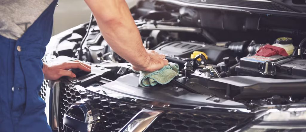 Auto technician wiping down the engine bay and front grille of a car with a microfiber cloth during routine vehicle inspection and maintenance in a repair shop.