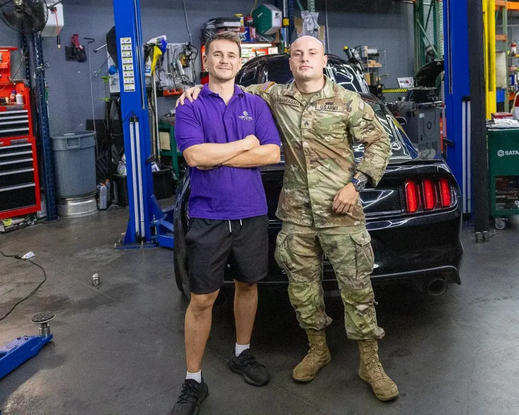A mechanic from Top Level Cars Hawaii shakes hands with a soldier. An auto repair shop in the background.